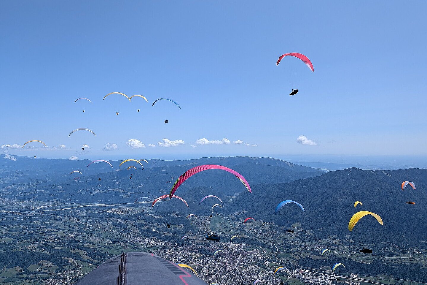 Viele Gleitschirme über einem Tal; unten ein Ort und Berge im Hintergrund