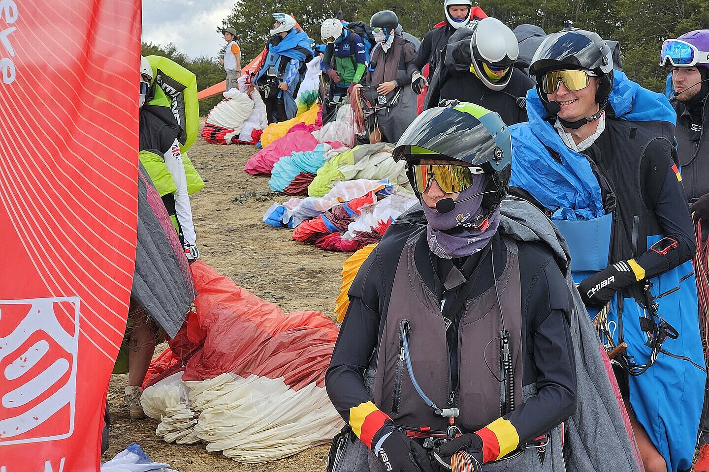 Ein Pilot wartet mit Gleitschirm am Startplatz, im Hintergrund liegen Schirme.