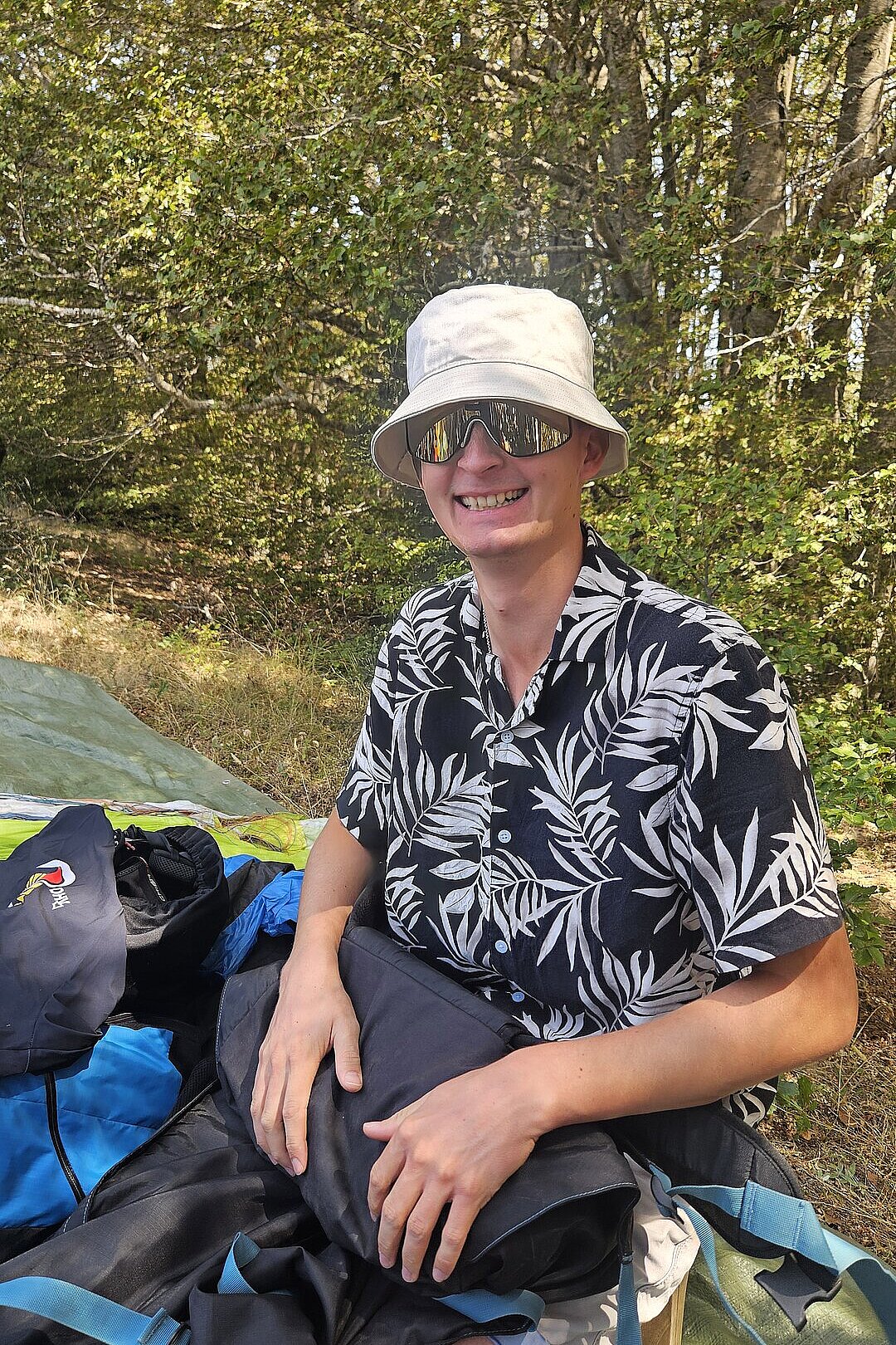 Person mit Sonnenhut und Sonnenbrille sitzt auf einer Decke im Wald und lächelt.