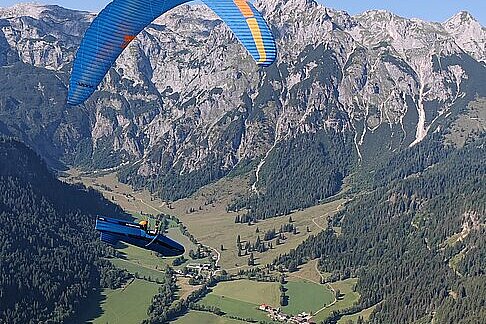 Zwei Gleitschirme über einem Tal mit Dörfern und hohen Bergen unter blauem Himmel.