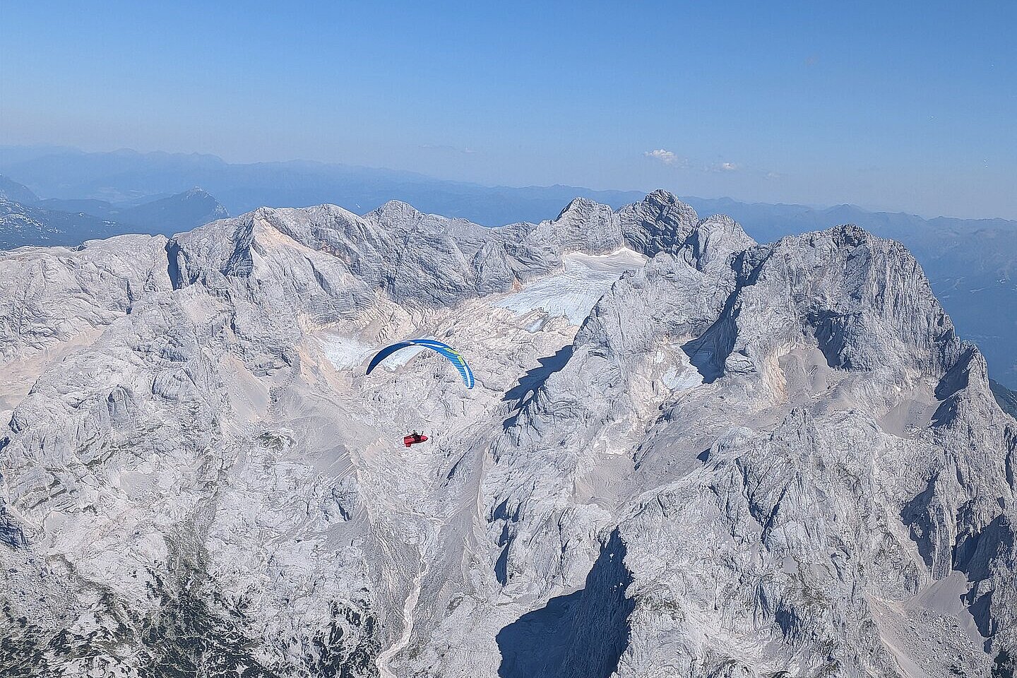 Ein Gleitschirm fliegt vor einer hellgrauen Felswand über einem Gebirgsmassiv.