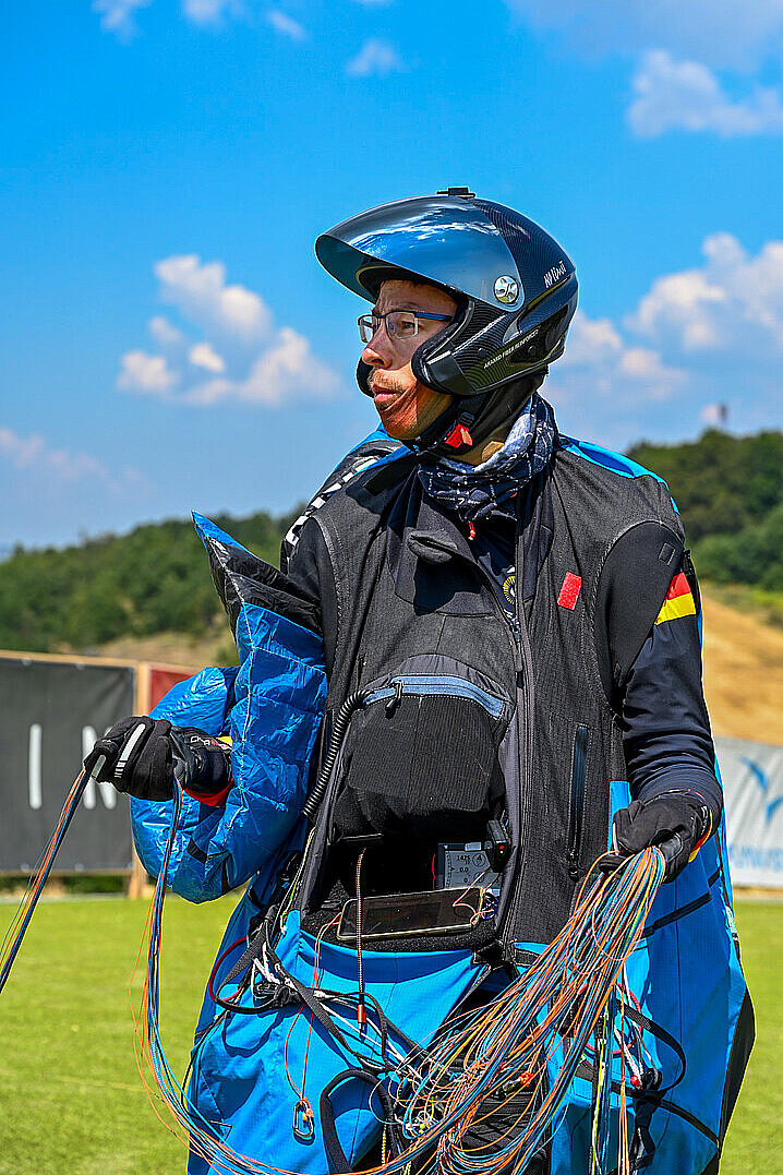 Pilot mit Helm und Flugsack steht auf einer Wiese und hält viele Leinen in den Händen.