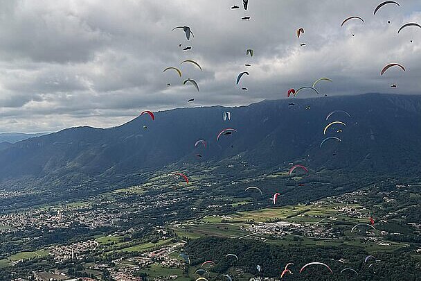 Viele Gleitschirme fliegen über ein Tal und Felder unter grauem Himmel.