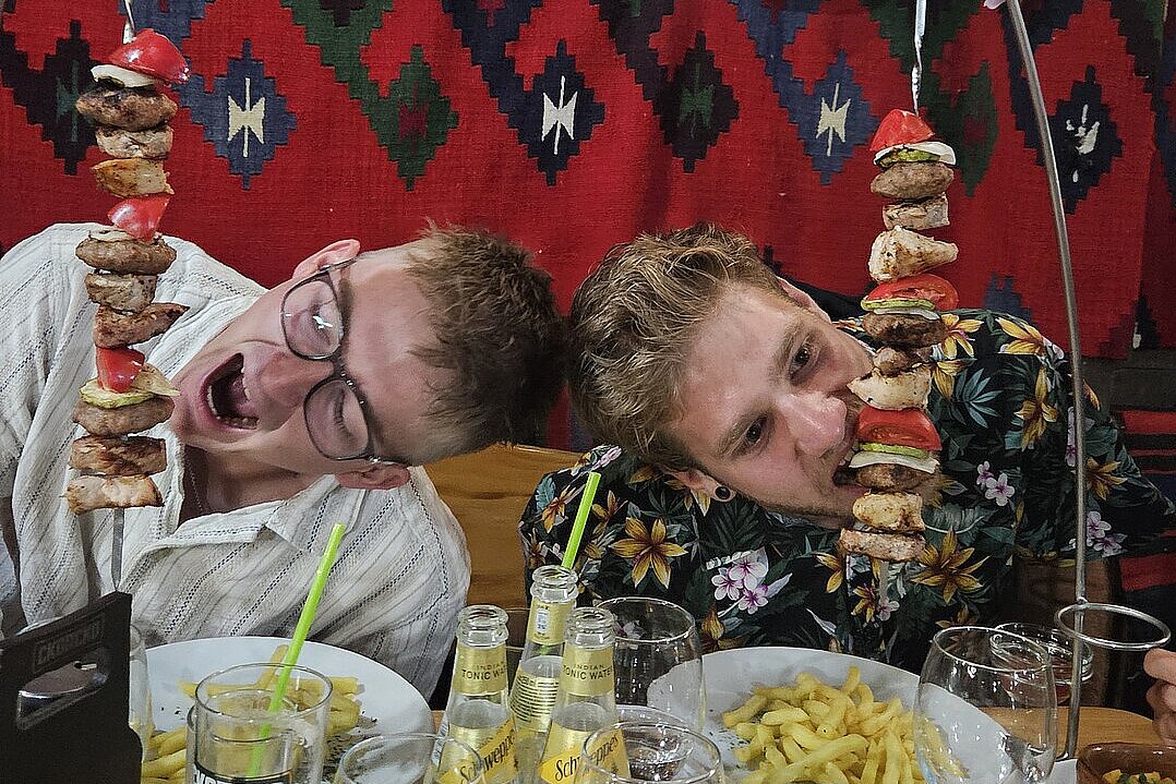 Zwei Männer am Tisch essen große Fleischspieße und Pommes.