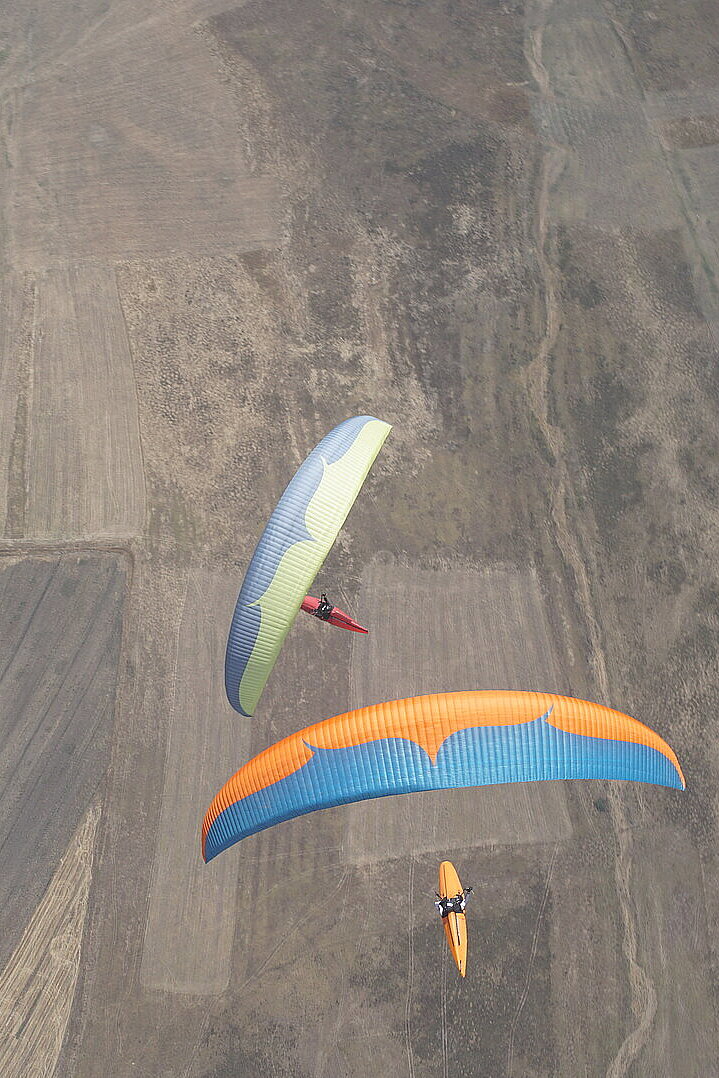 Mehrere Gleitschirme fliegen über Felder in der Nähe eines Ortes.