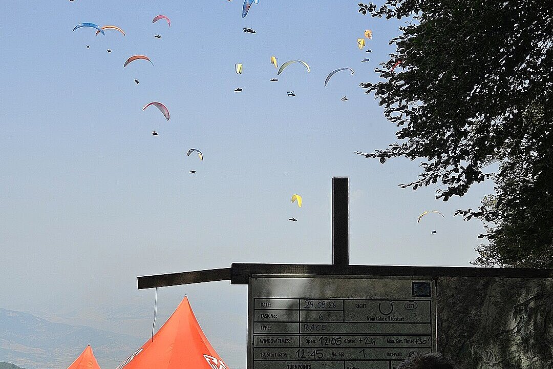 Pilotengebiet am Startplatz mit Tasktafel, roten Pavillons und mehreren Gleitschirmen am Himmel.