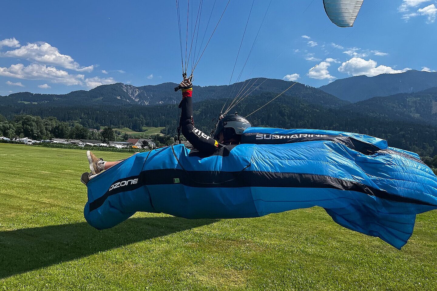 Ein Pilot sitzt im Liegegurtzeug über einem grünen Feld, darüber ist ein Flügel zu sehen.