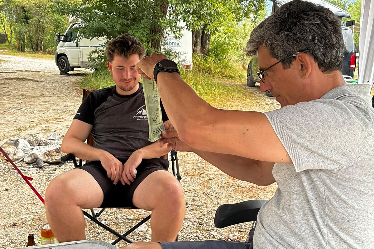 Unter einem Zelt wird ein sitzender Mann von einem anderen mit einem Beutel versorgt.