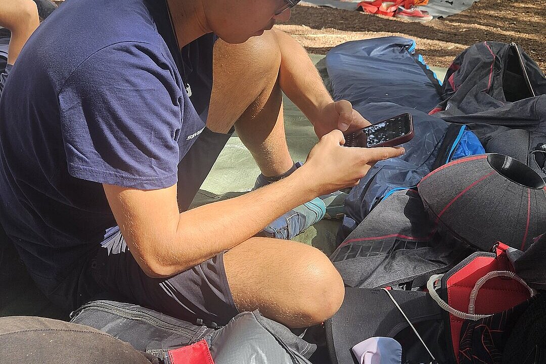 Eine Person sitzt im Schatten und schaut auf ein Smartphone, daneben liegen Gleitschirm-Ausrüstungen.
