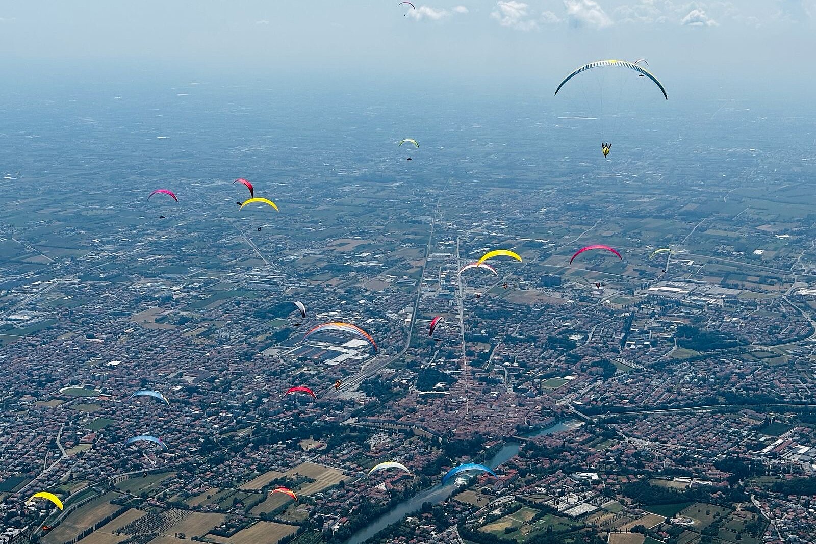 Viele Gleitschirme über einer Stadt und Feldern, weit am Himmel verteilt.