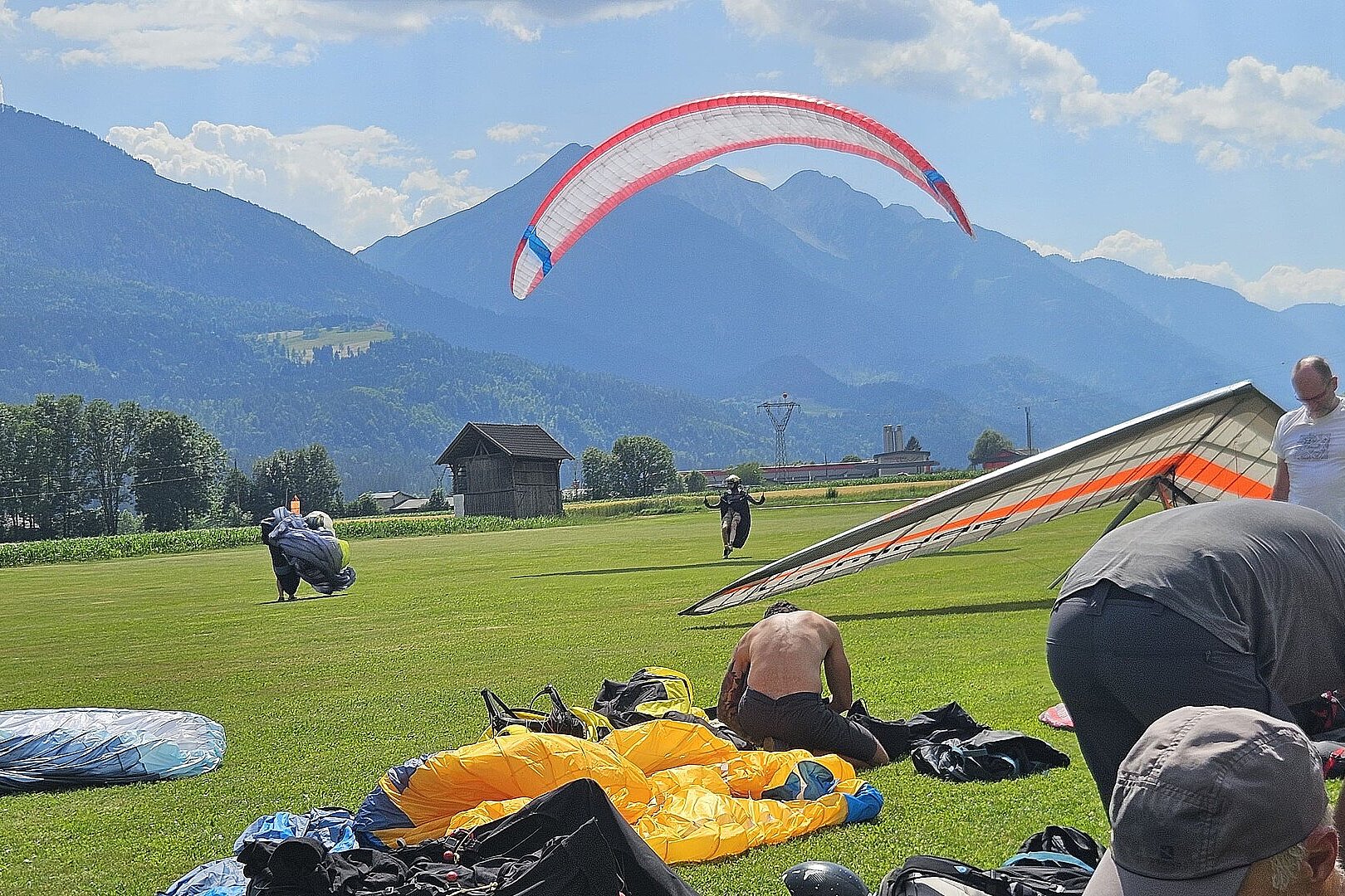 Gleitschirm am Himmel, daneben andere Schirme und Personen beim Aufbauen auf einer Wiese.