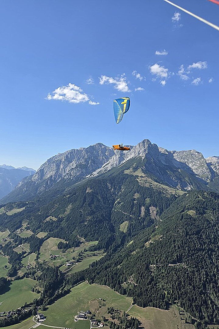 Gleitschirm mit blau-gelbem Schirm fliegt vor Bergkulisse über grüne Täler.