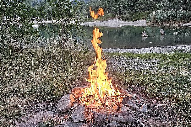 Lagerfeuer am Ufer vor einem See, dahinter ein hoher Berg und Bäume.