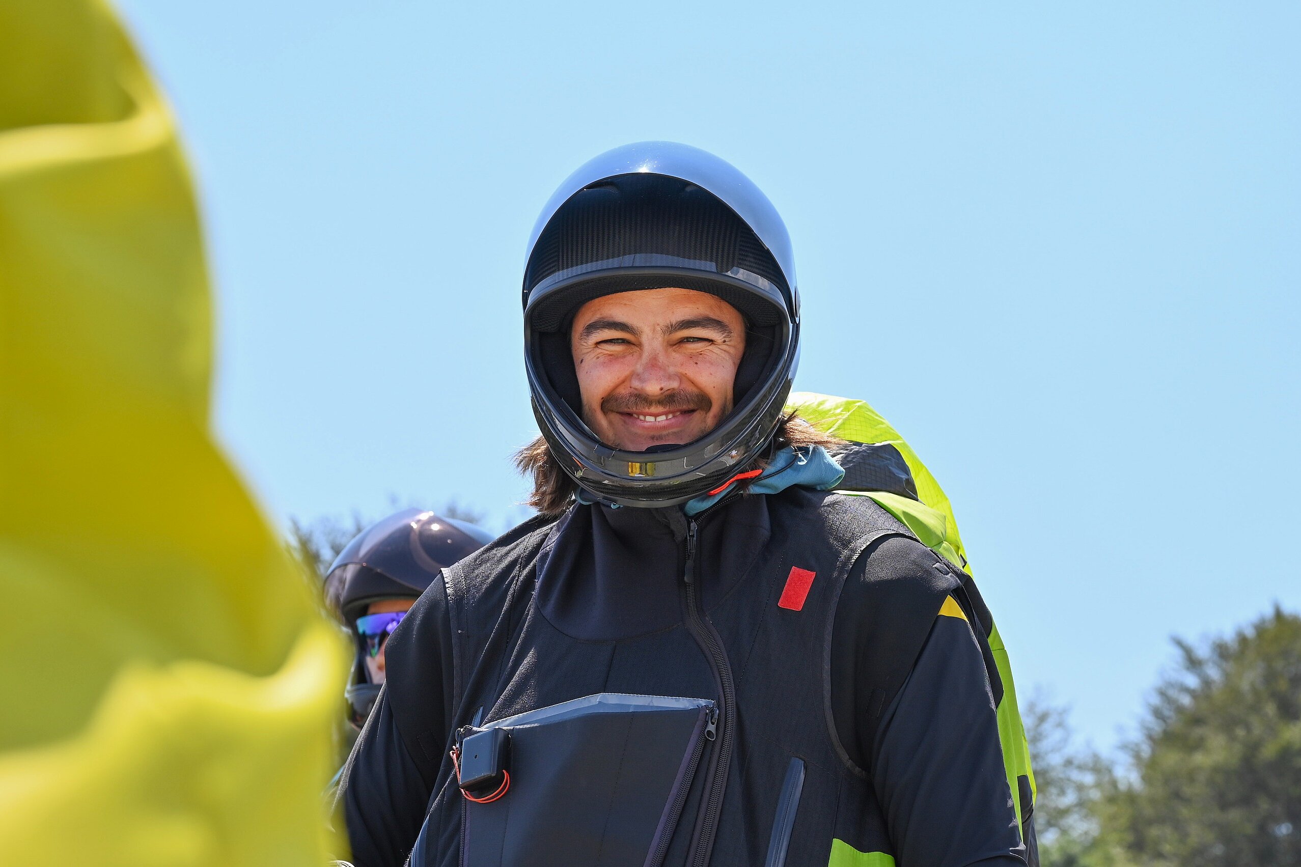 Pilot in schwarzem Anzug und Helm lächelt am Startplatz in die Kamera.