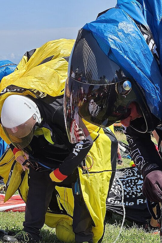 Pilotin in Startvorbereitung mit Helm, Gurtzeug und ausgebreiteten Schirmen auf der Wiese