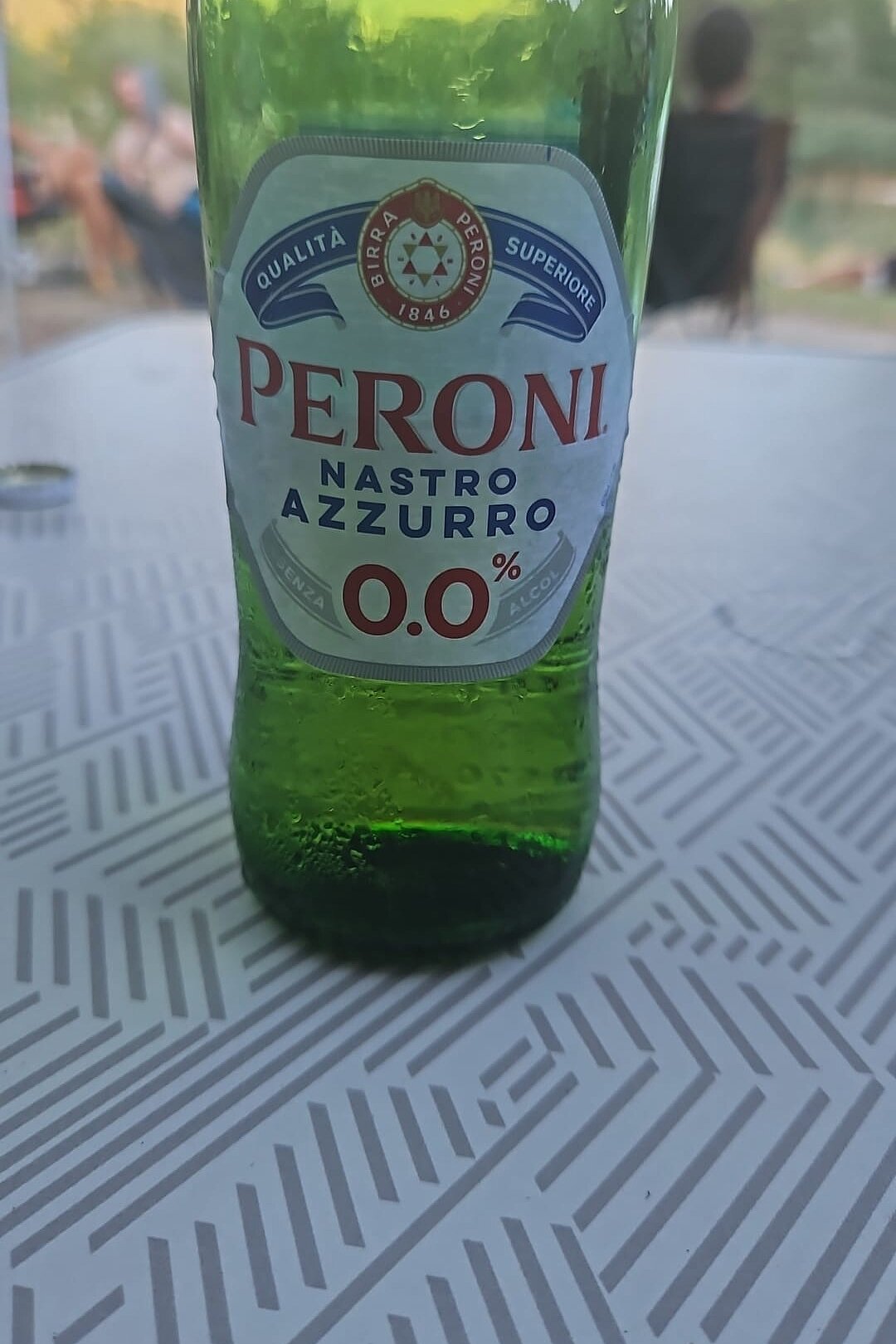 Grüne Peroni-Nastro-Azzurro-0,0-Flasche steht auf einem Tisch, im Hintergrund unscharf sitzende Personen.