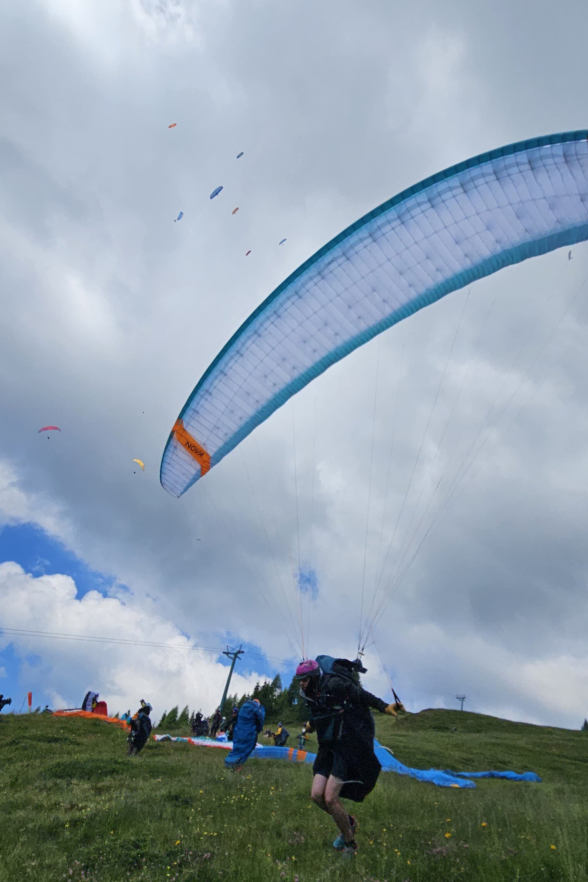 Pilot startet mit aufgespanntem Gleitschirm auf einer Wiese, weitere Schirme und Personen im Hintergrund.