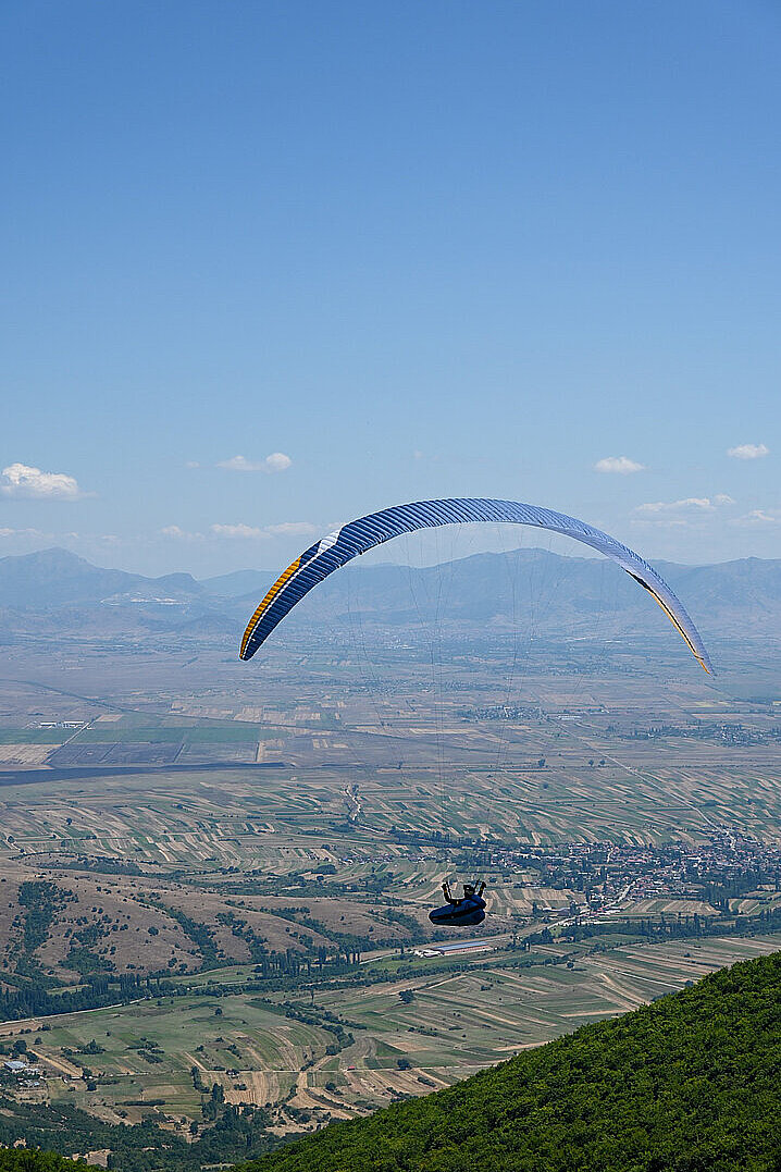 Ein Gleitschirmflieger schwebt über einer Landschaft mit Feldern und Hügeln.