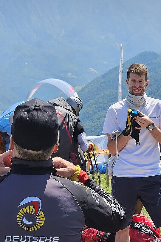 Mehrere Piloten bereiten am Startplatz ihre Ausrüstung vor, Berge im Hintergrund