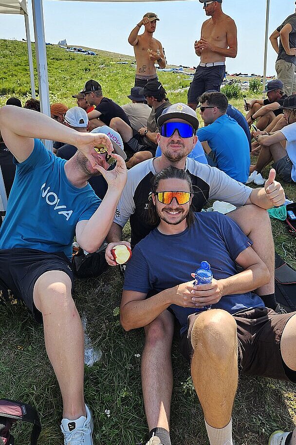Mehrere Männer sitzen und stehen auf einer Wiese unter einem Pavillon, einige mit Sonnenbrillen und Getränken.
