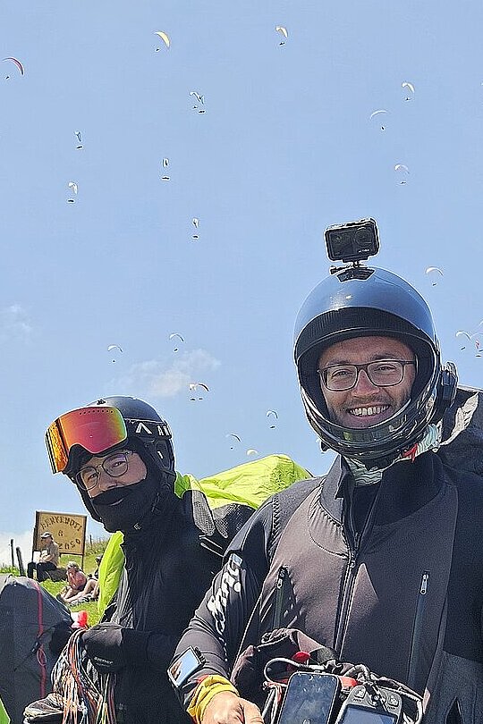 Zwei Piloten mit Helm und Packtaschen lächeln am Startplatz, viele Gleitschirme steigen im Himmel