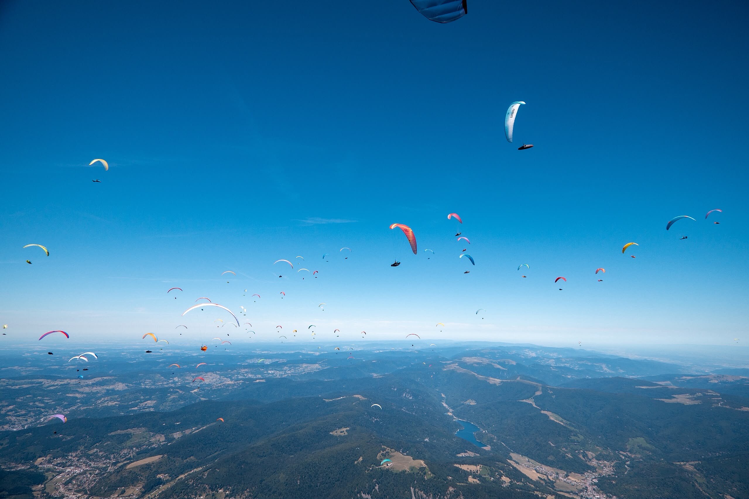 Viele Gleitschirmflieger schweben über einer Berglandschaft unter klarem blauem Himmel.