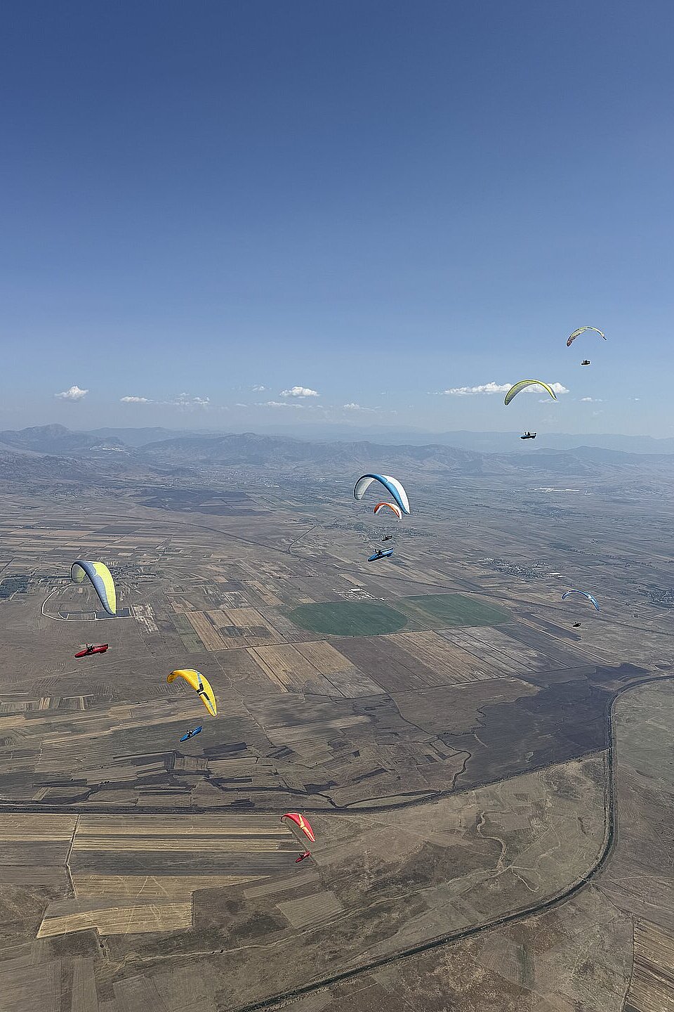 Mehrere Gleitschirme fliegen über einer weiten, kahlen Landschaft mit Feldern und Bergen im Hintergrund.