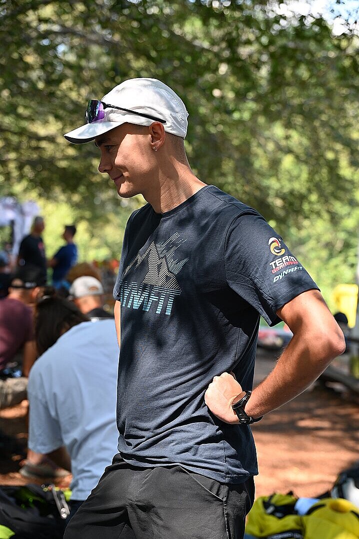 Ein junger Mann im Team-Shirt steht mit verschränkter Haltung zwischen anderen Piloten im Schatten.