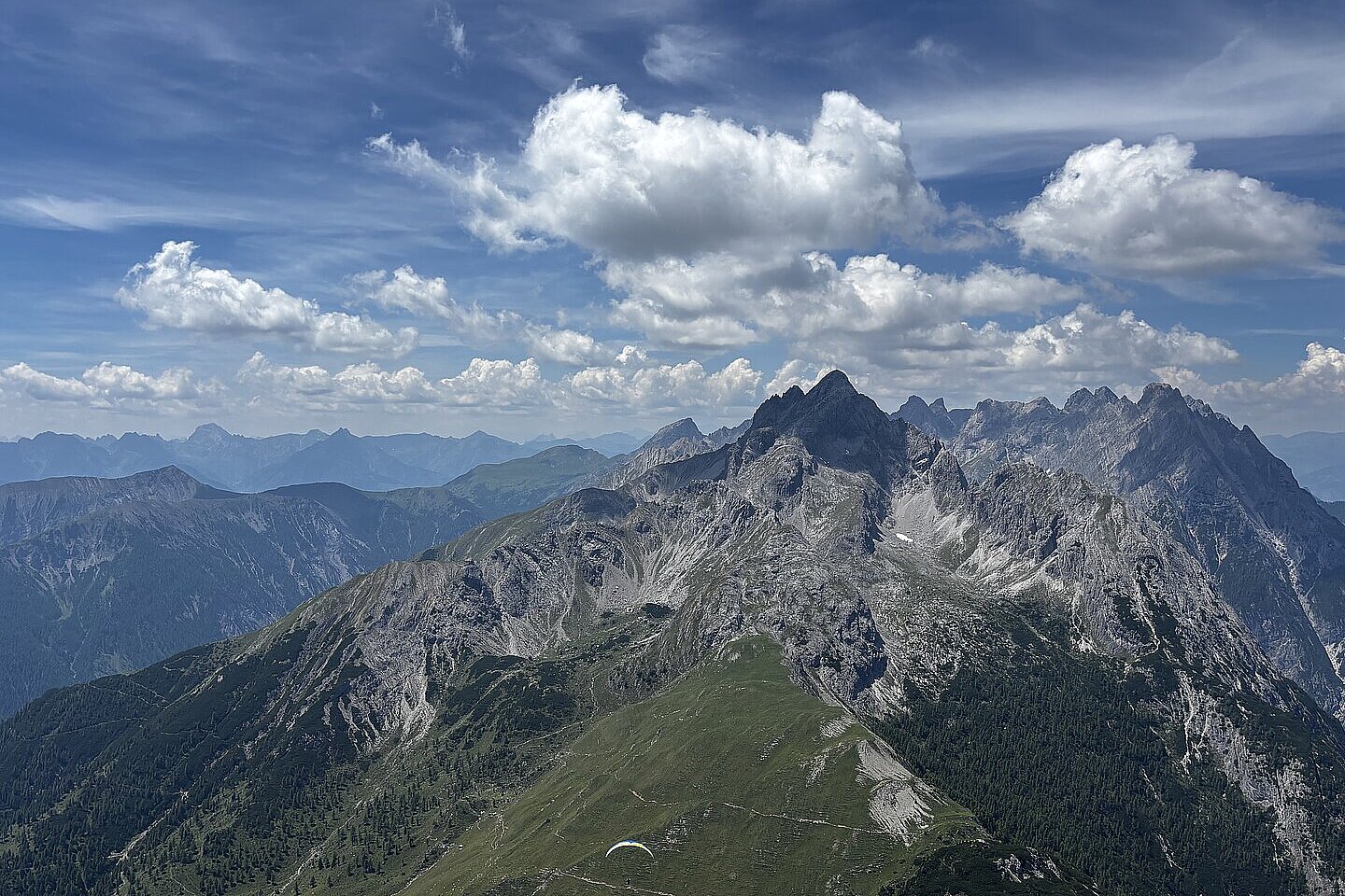Bergkamm mit Felsgipfeln unter wolkigem Himmel; unten schwebt ein einzelner Gleitschirm.