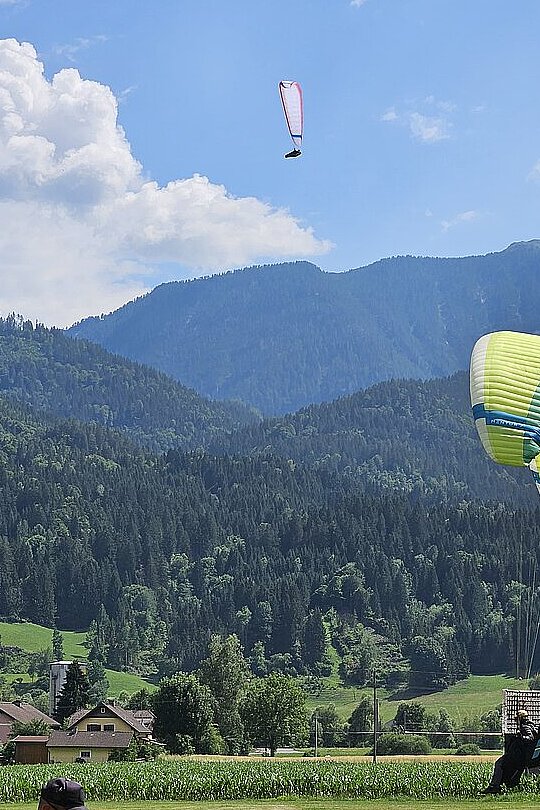 Zwei Gleitschirme fliegen über einer grünen Wiese vor Bergen und Wolken. Ein Mann steht unten.