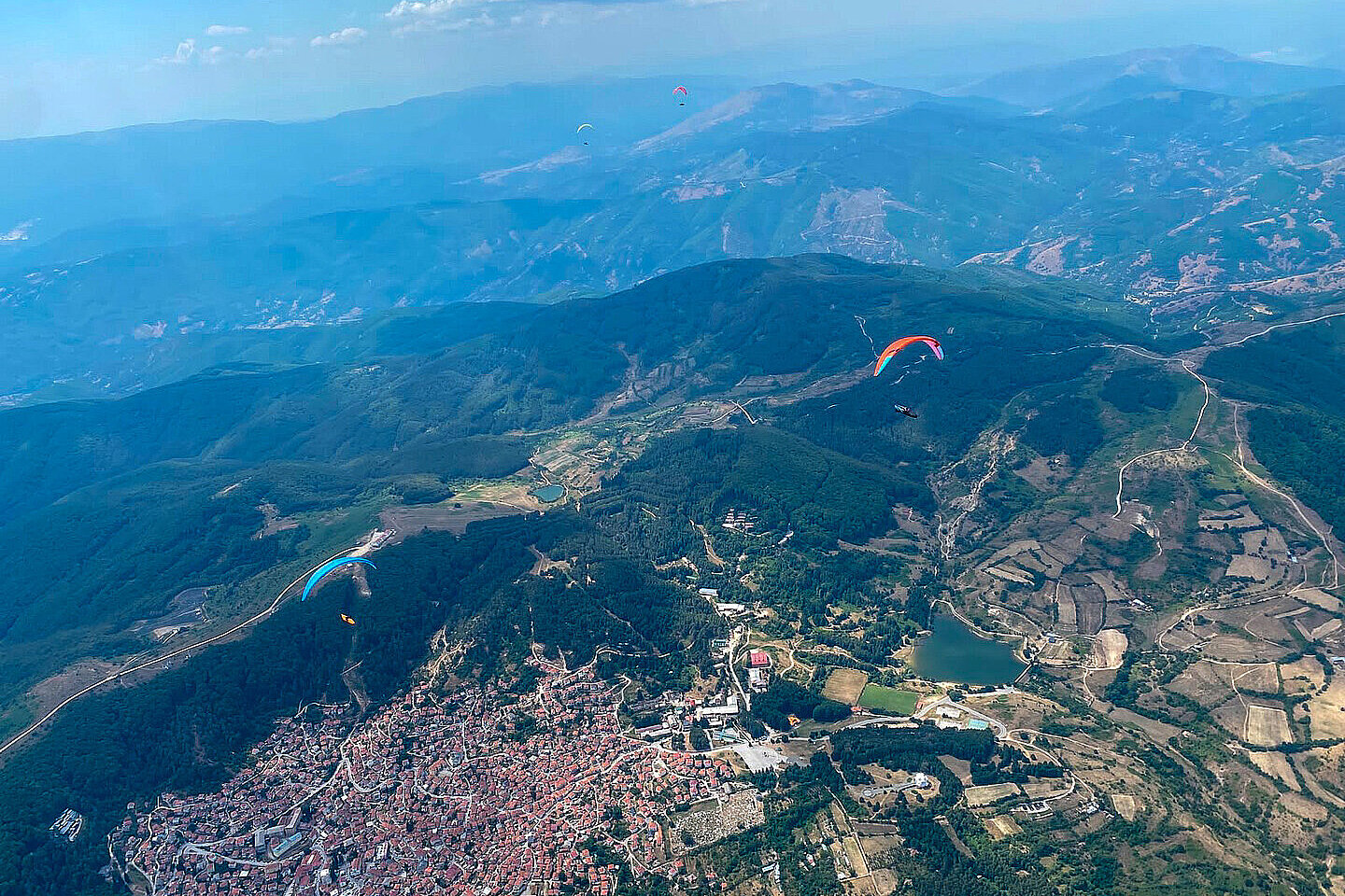 Mehrere Gleitschirme fliegen über eine bergige Landschaft mit einer Stadt und Feldern darunter.