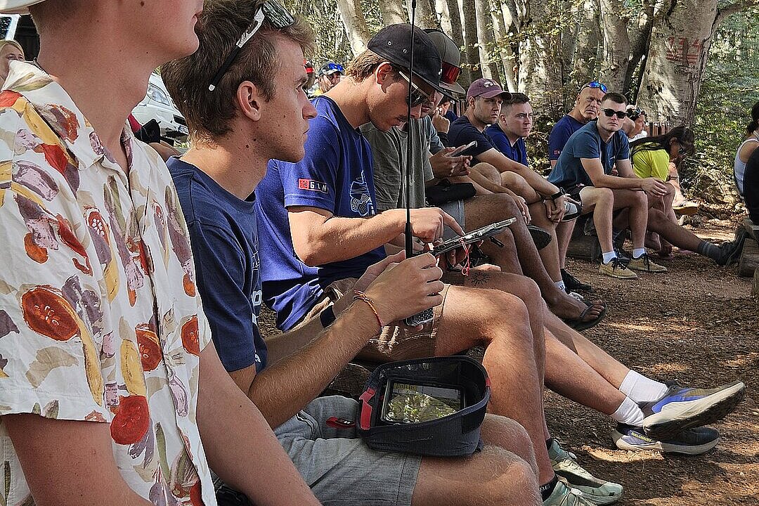 Junge Männer sitzen im Schatten auf einer Bank und warten, einige halten Funkgeräte oder Smartphones.