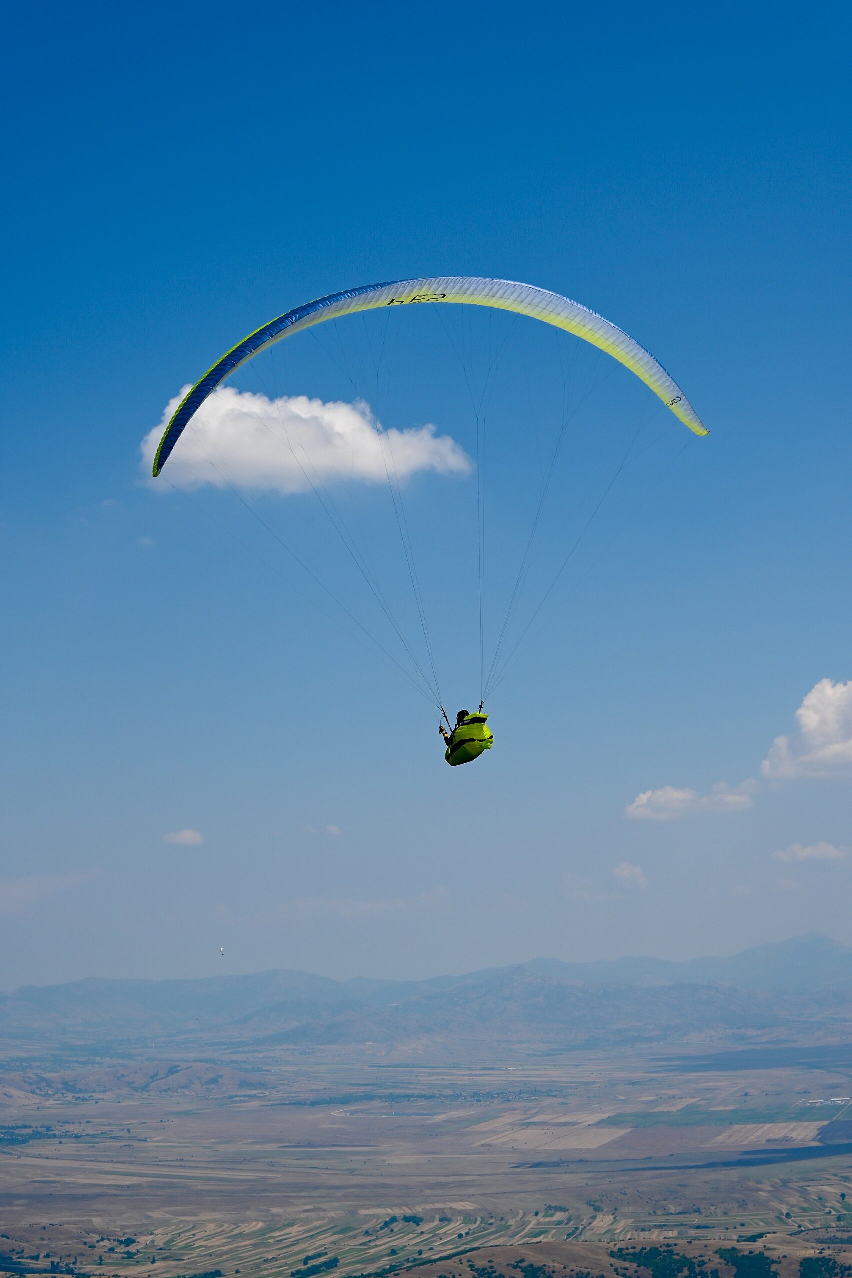 Gleitschirm mit gelb-grünem Schirm fliegt vor blauem Himmel über einer Ebene.