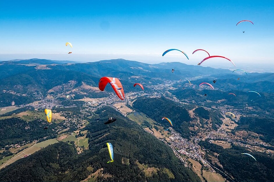 Viele Gleitschirme schweben über bewaldeten Hügeln und einem Tal.