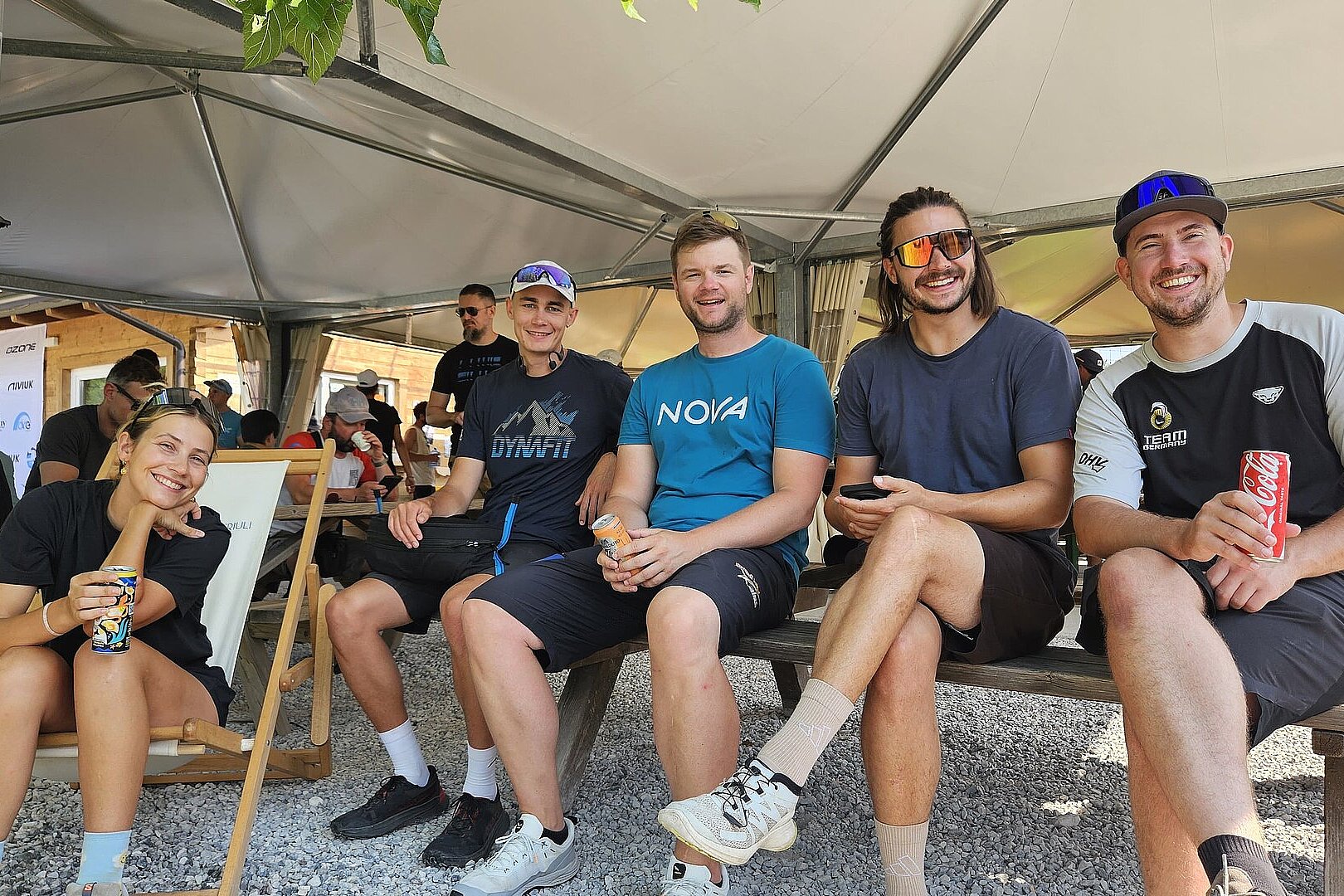Gruppe von sechs Personen sitzt lachend unter einem Pavillon und hält Getränke in der Hand.