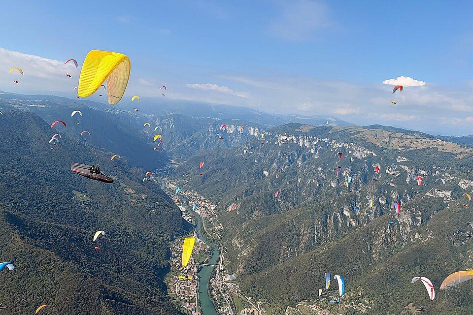 Mehrere Gleitschirme fliegen über ein Tal mit Fluss, Bergen und verstreuten Orten.