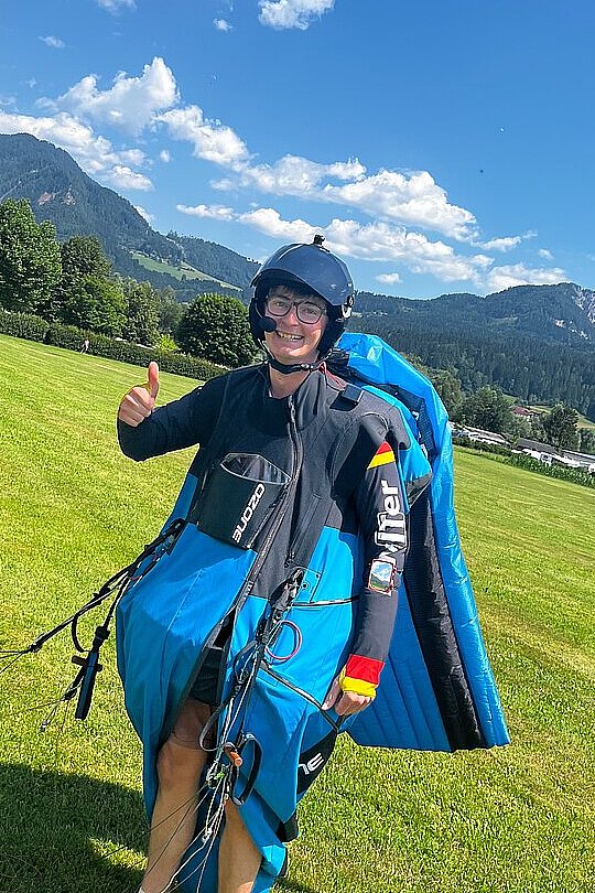 Lächelnde Person im Gleitschirm-Gurtzeug auf einer Wiese, mit Daumen hoch vor Bergkulisse.