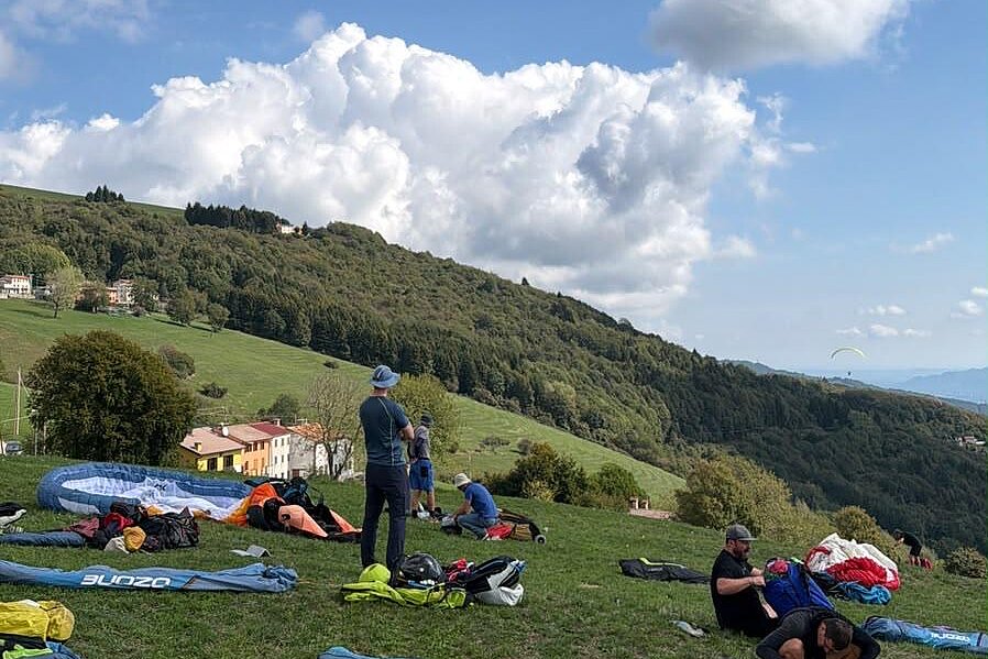 Mehrere Personen packen auf einer Wiese Gleitschirmausrüstung zusammen, während am Himmel Gleitschirme fliegen.