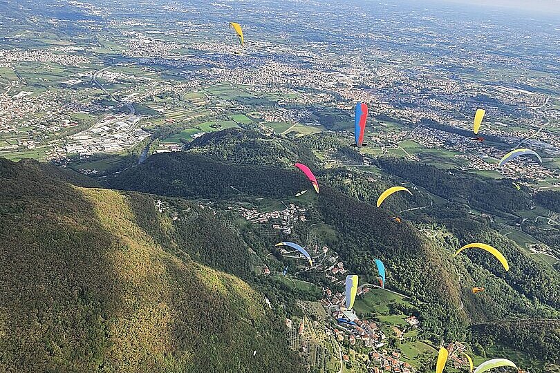 Viele Gleitschirme fliegen über grün bewaldeten Hügeln und einer weitläufigen Stadtlandschaft.