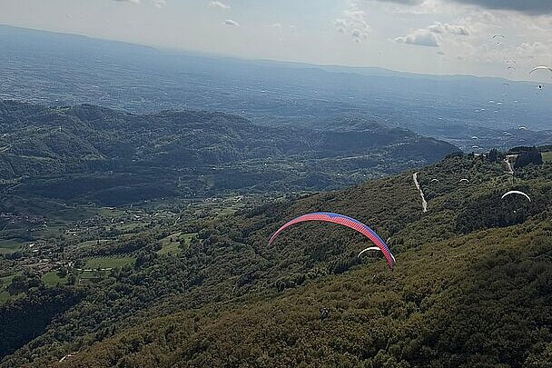 Ein Gleitschirm fliegt über bewaldete Hänge und ein Tal unter blauem Himmel.