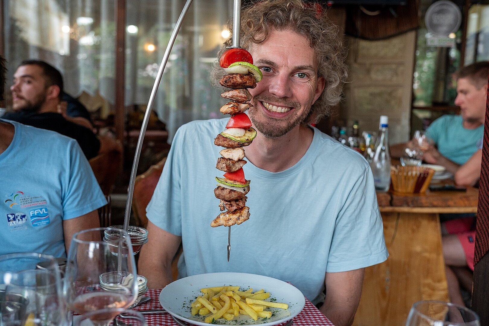 Mann sitzt am Tisch vor einem Teller Pommes und einem vertikalen Grillspieß mit Fleisch und Gemüse.
