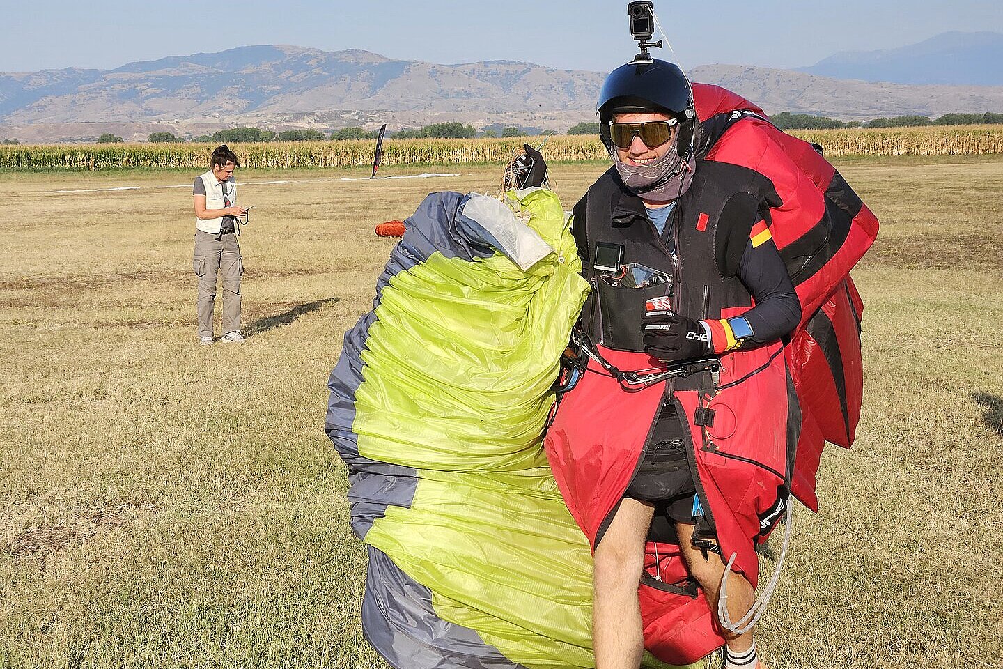 Pilot mit rotem Gleitschirmrucksack steht auf einer Wiese, ein weiterer Mensch schaut im Hintergrund aufs Handy.