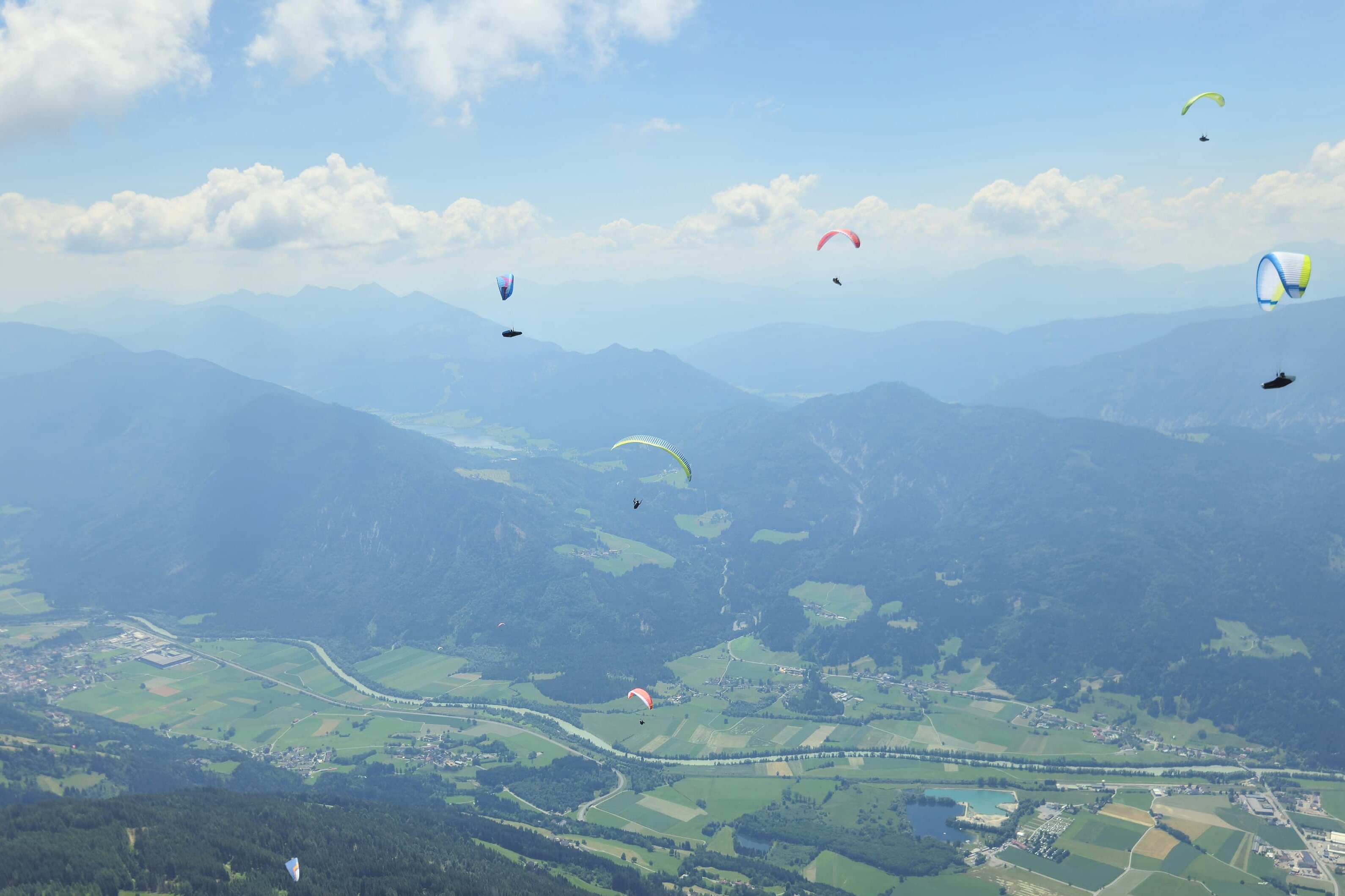 Mehrere Gleitschirme schweben über einem breiten Tal mit Feldern und Bergen im Hintergrund.