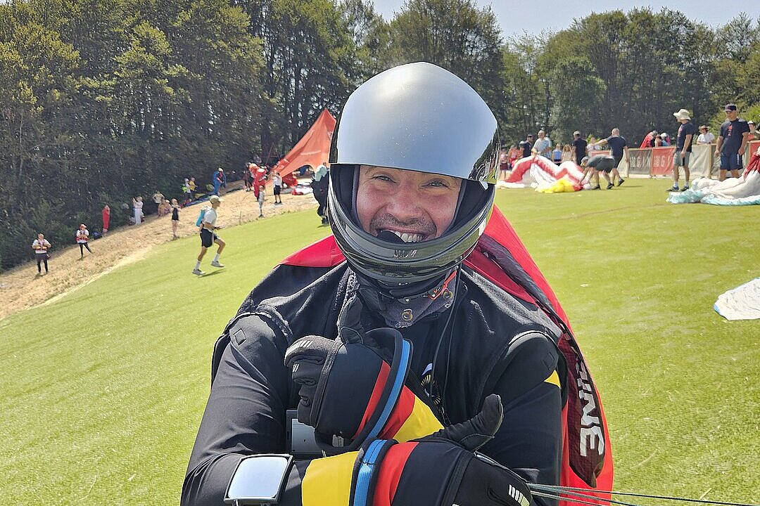 Ein Pilot im Helm und Gurtzeug lächelt auf einem grünen Startplatz und hält die Leinen fest.
