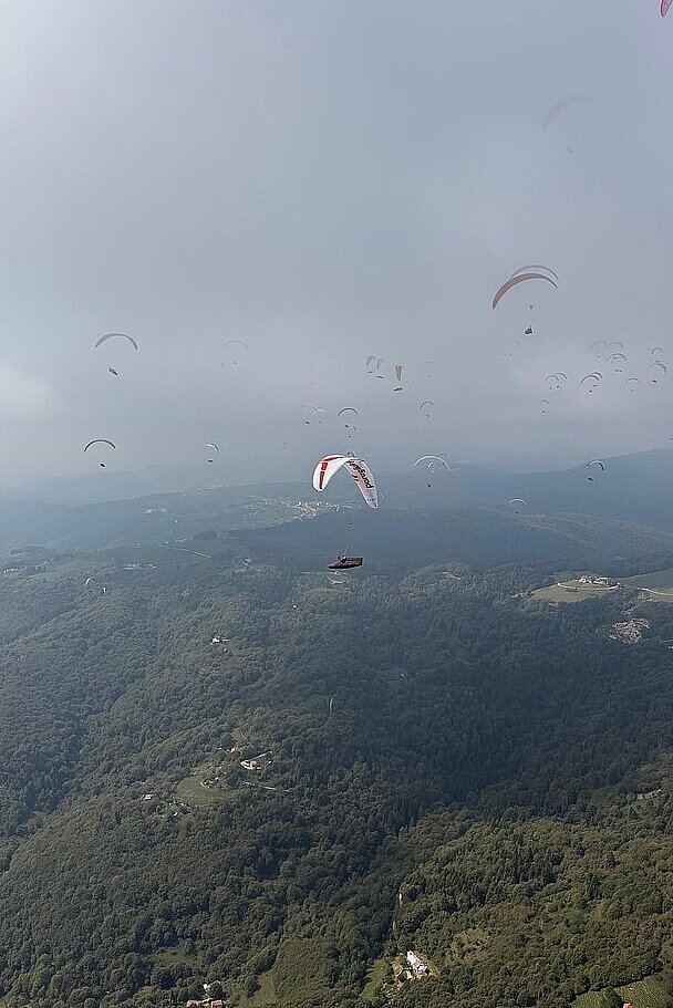 Viele Gleitschirme über bewaldeten Hügeln unter trübem Himmel.