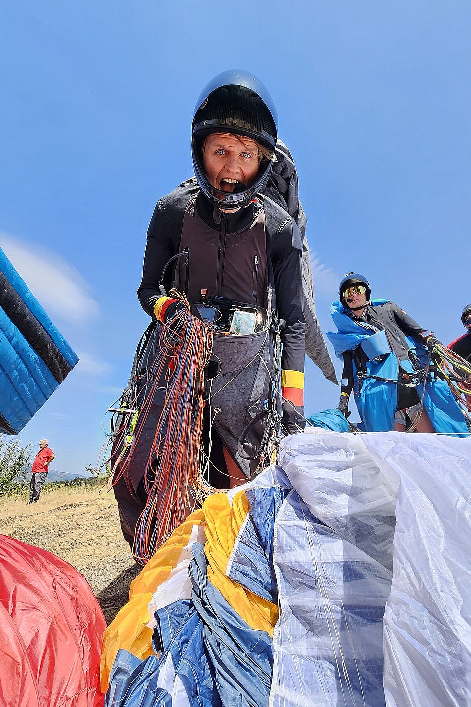 Pilotin mit Helm hält Leinen und schaut nach vorn, andere bereiten Schirme auf der Wiese vor.