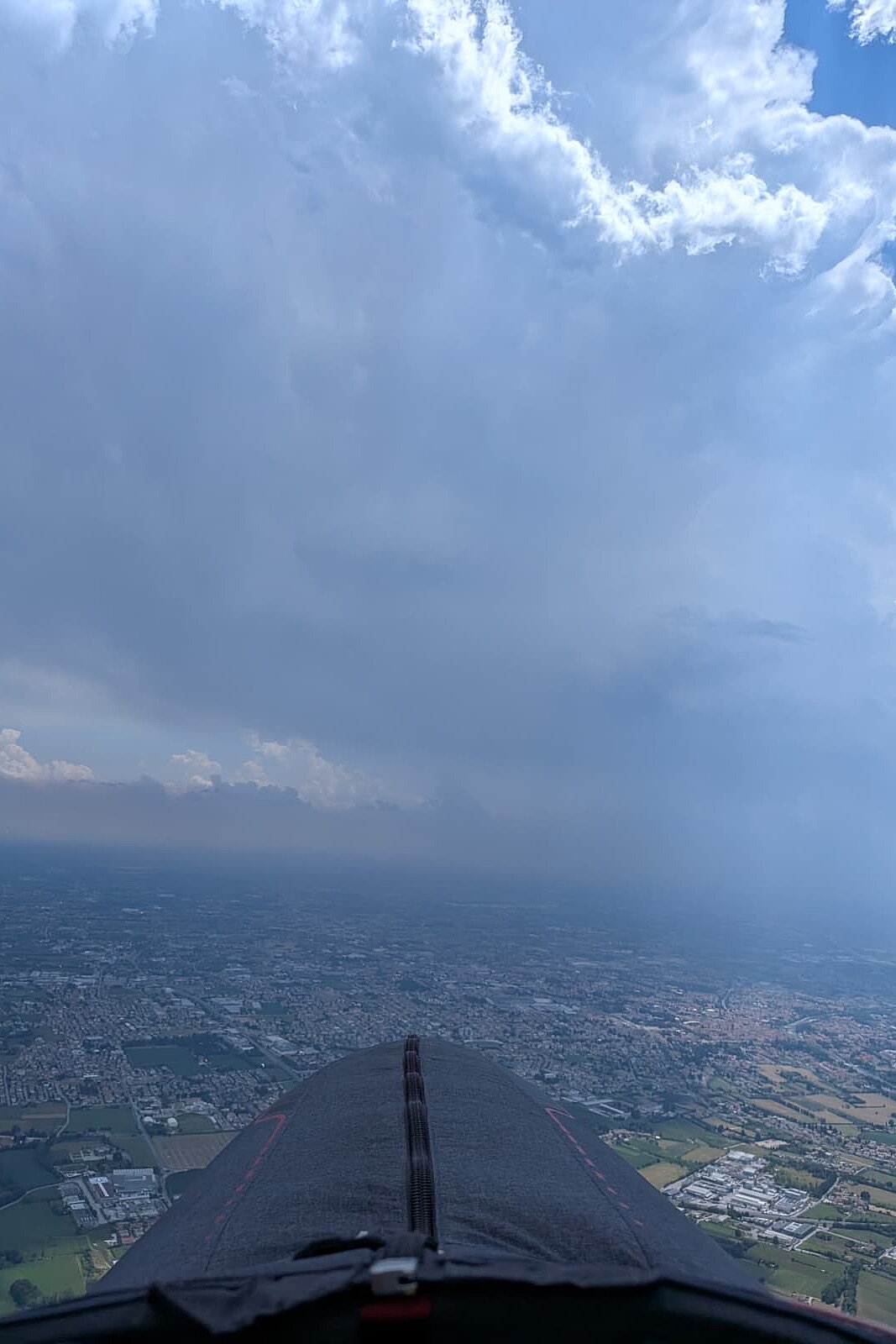 Blick aus dem Gleitschirm über eine Stadt und ein Gewitterfeld am Horizont.