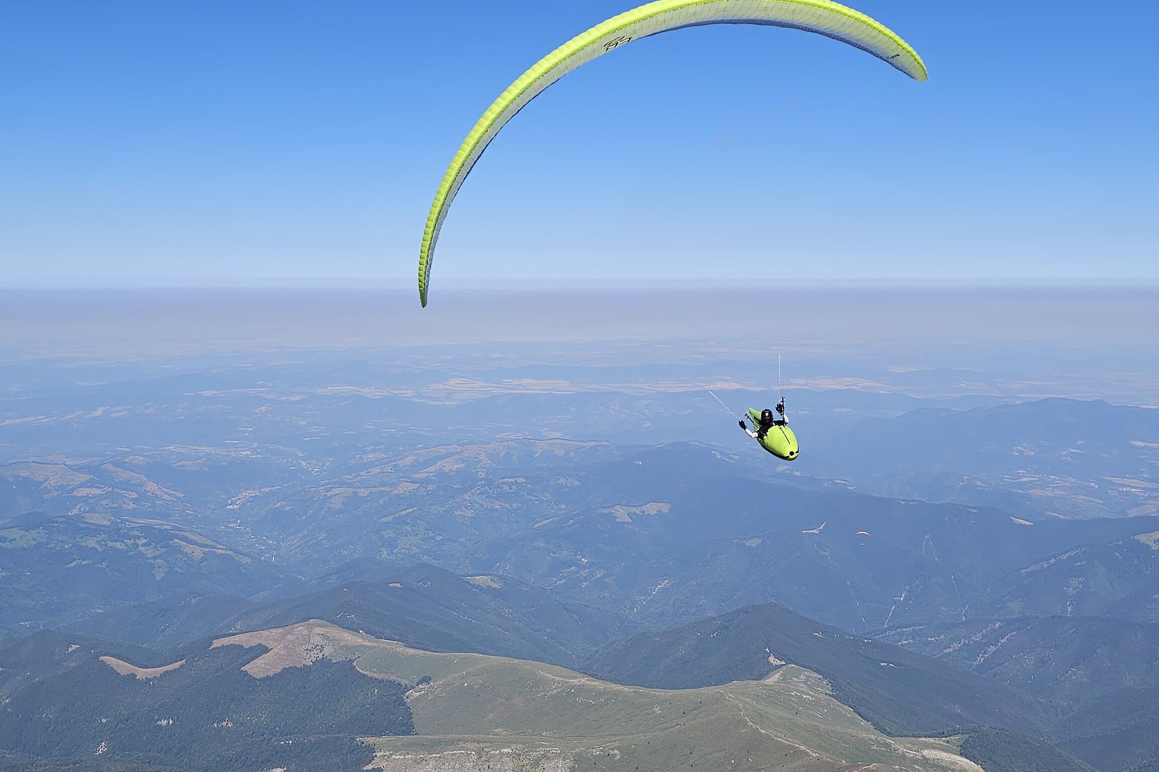 Ein Gleitschirmpilot fliegt über Gebirgslandschaft und weite Täler unter klarem Himmel.