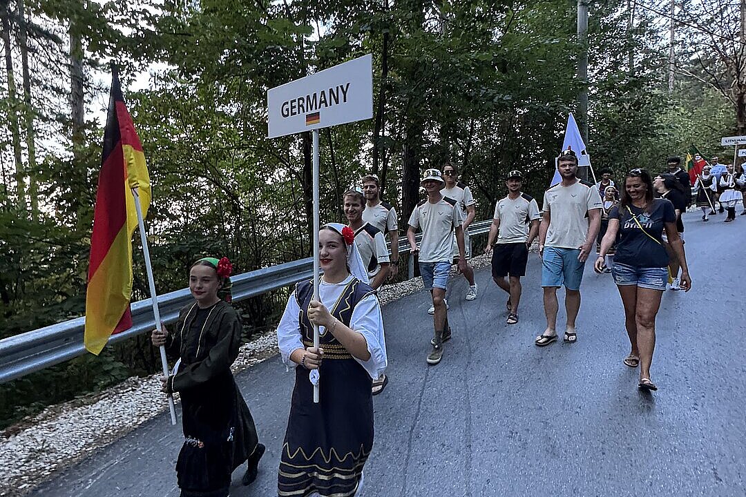Eine Gruppe marschiert mit Deutschland-Schild und Flaggen auf einer Straße durch den Wald.