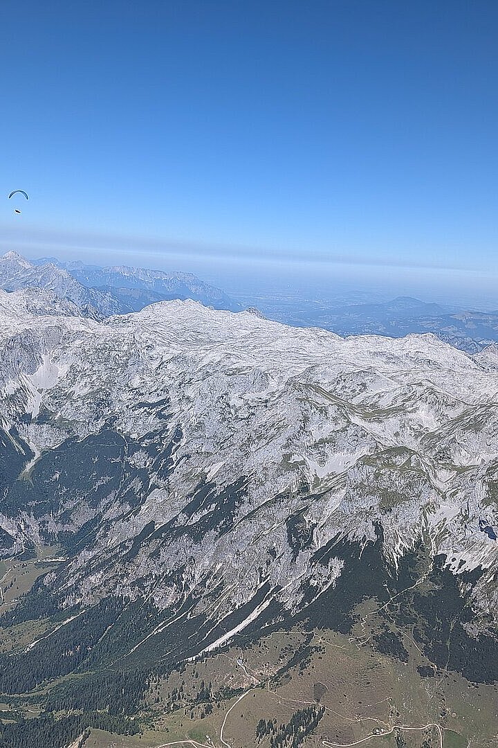 Schneebedeckte Bergkette unter blauem Himmel, ein einzelner Gleitschirm weit oben links.